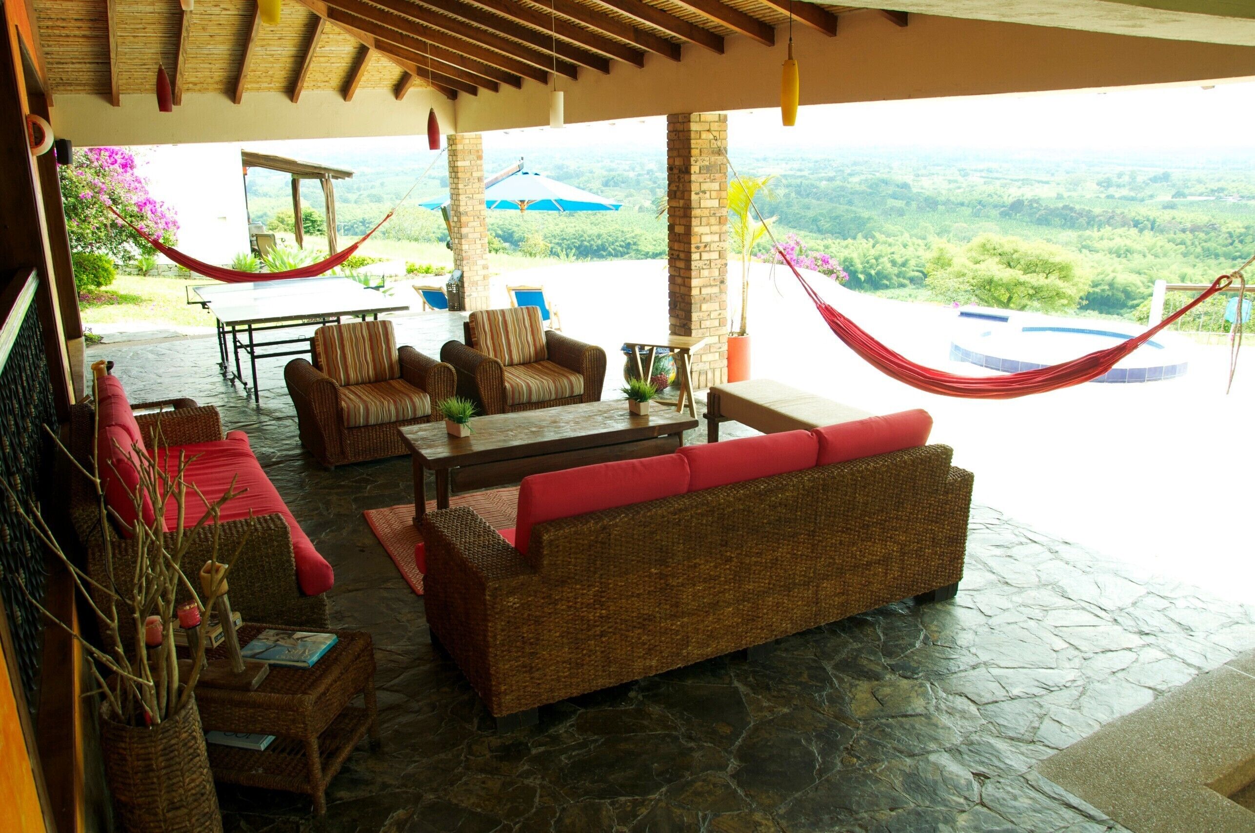 A covered outdoor seating area with wicker furniture, a stone floor, and a view of the landscape.