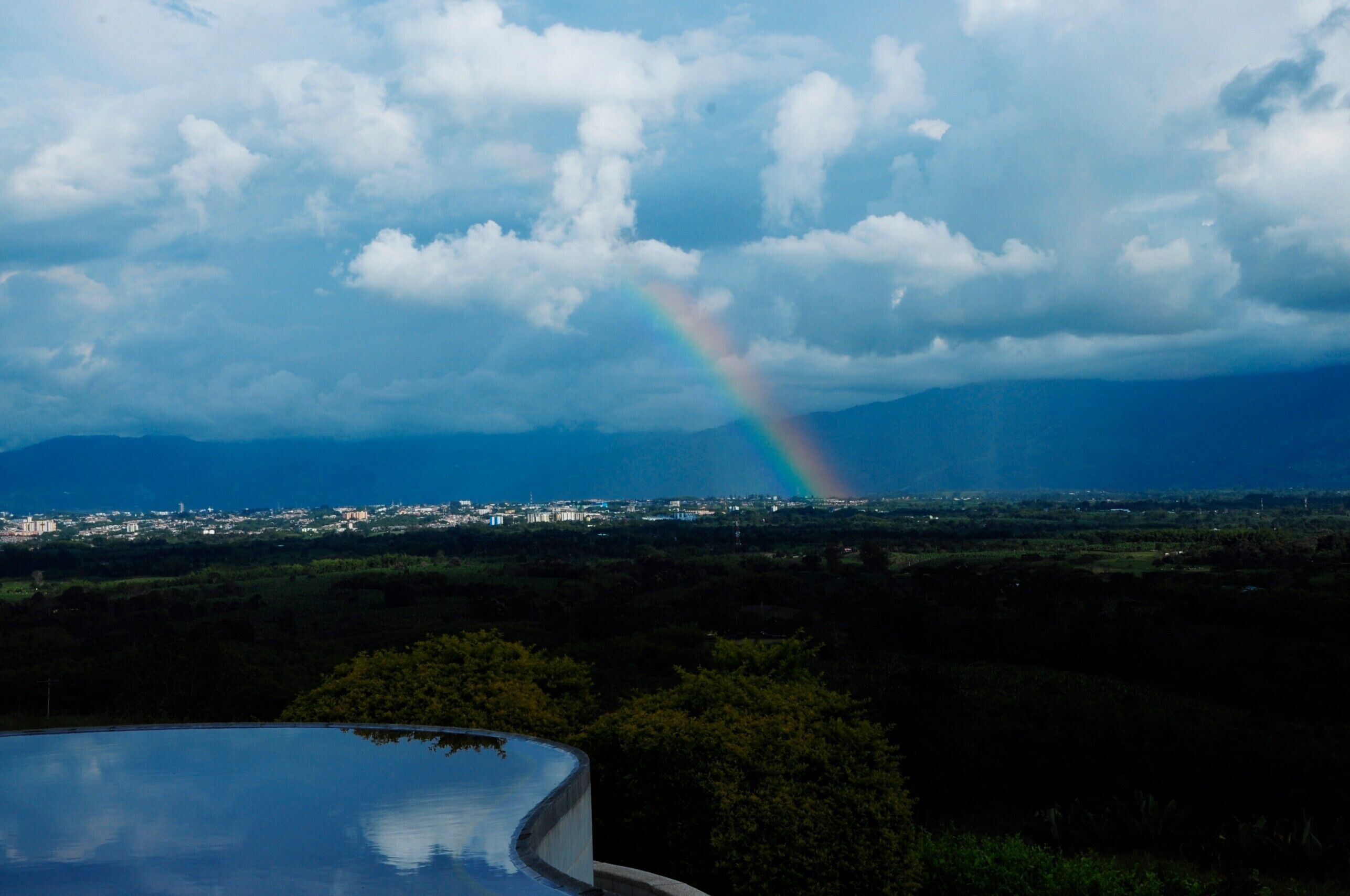 A cityscape with a rainbow, mountains in the background, and a body of water in the foreground.