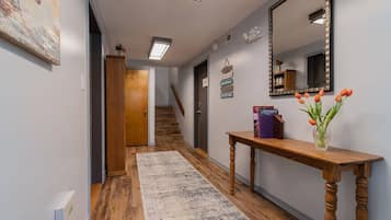 A hallway with a wooden floor, a rug, a wooden console table with a vase of flowers, a mirror, and a staircase in the background.