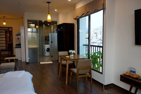 A hotel room with a bed, a dining table with chairs, a small kitchen area, and a balcony with a view of the city.