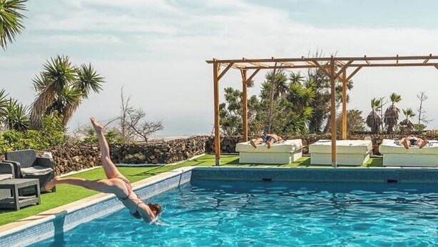 Une piscine dans laquelle une personne plonge, des chaises longues et une pergola où des gens se détendent.
