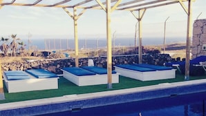 Un espace piscine avec des chaises longues disposées sous une pergola, un mur en pierre et une vue sur l’océan.