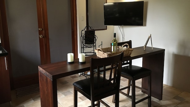 Un comedor con una mesa de madera, dos sillas, un televisor montado en la pared y una botella de vino sobre la mesa.