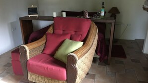 Un sillón de mimbre con cojines rojos, una mesita de madera con una botella de vino y una copa, y una pizarra con un mensaje de bienvenida.
