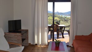 Ein Zimmer mit Fernseher auf einem Backsteinbock, einem Tisch mit Pflanze, einer Couch und einem Balkon mit Tisch und Stühlen mit Blick auf eine reizvolle Landschaft.