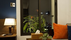 Une chambre avec une table de chevet, une lampe, une plante, une boîte de mouchoirs et une fenêtre donnant sur la ville la nuit.