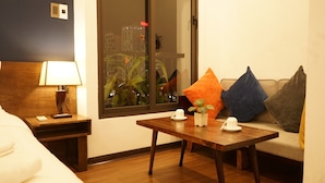 Une chambre d’hôtel avec une table de chevet en bois, une table de nuit avec une lampe, un canapé agrémenté de coussins colorés, une petite table avec une plante et une tasse, et une fenêtre donnant sur des immeubles.