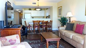 A living room with a sofa, a coffee table, and a TV. There is a kitchen in the background with a dining table and chairs. - Grand Atlantic by Palmetto Vacations (Myrtle Beach)