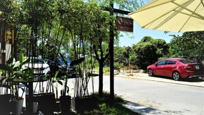 Vista da estrada do Hotel Oasis, com uma placa, plantas em vasos e um carro vermelho estacionado nas proximidades.