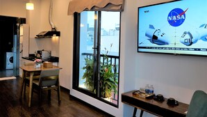Quarto de hotel moderno com varanda, televisão exibindo veículos espaciais da NASA, área de refeições com mesa e cadeiras, e pequena cozinha.