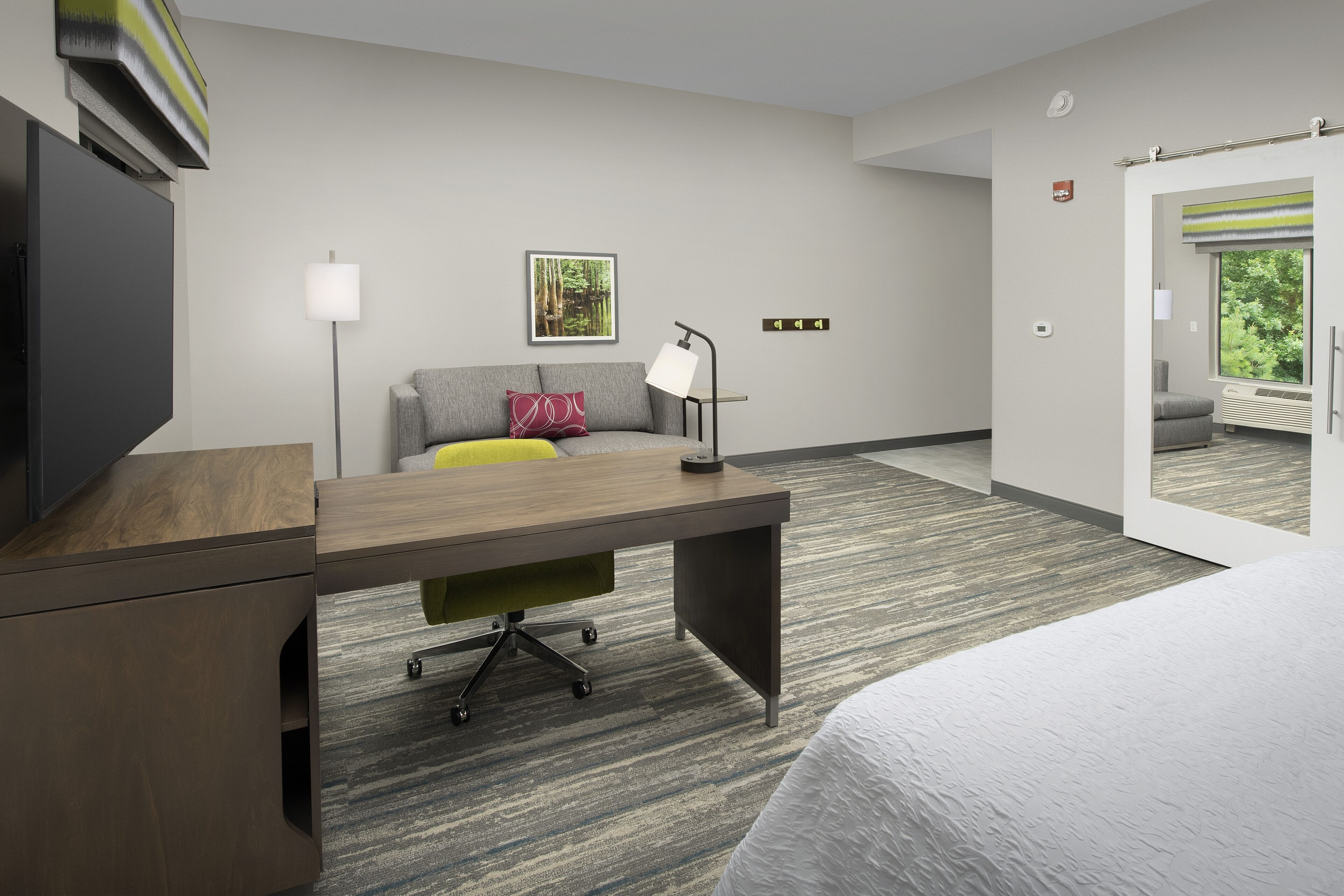 Quarto de hotel moderno com escrivaninha de madeira, sofá cinzento, cama, abajur e quadro na parede.