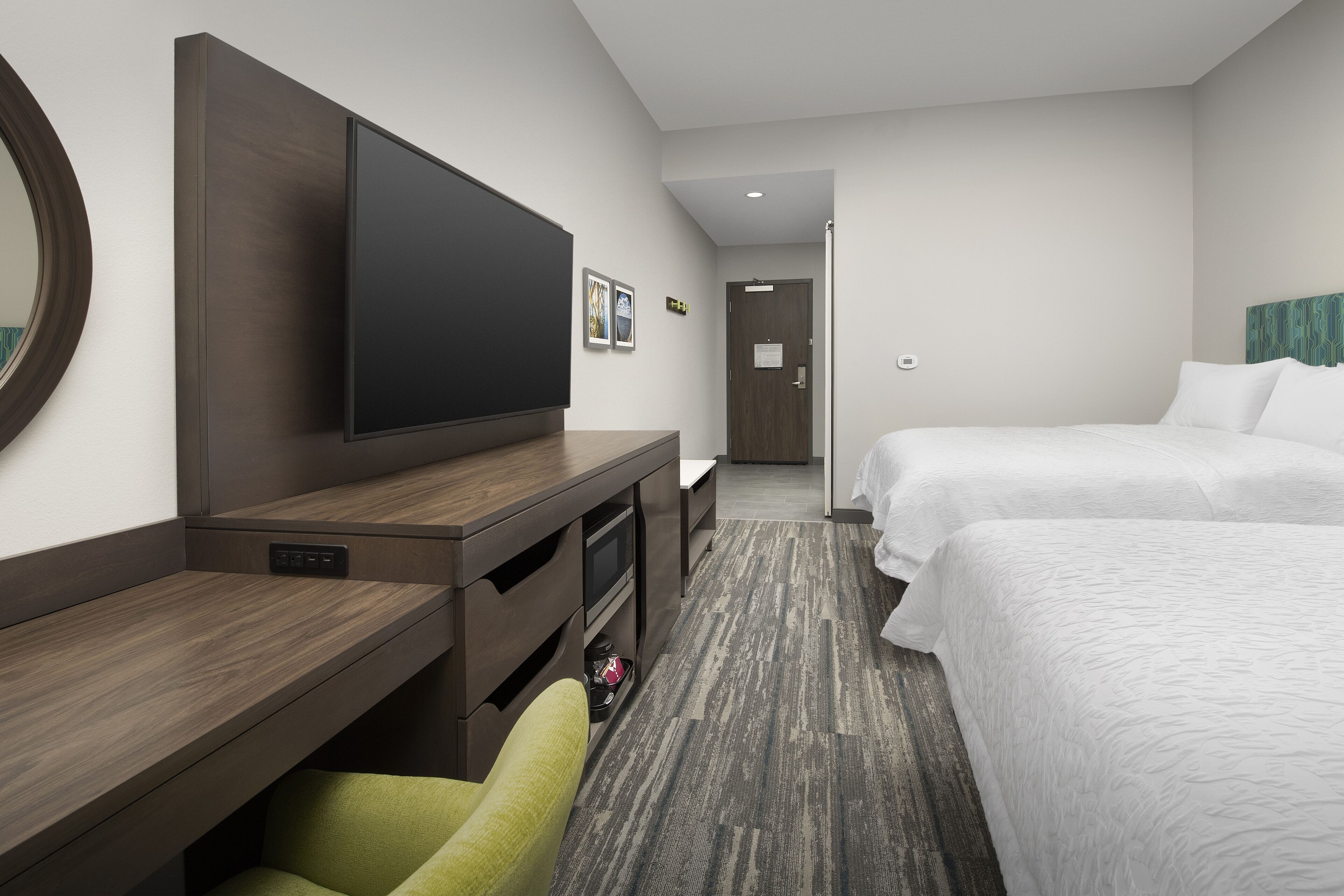 A hotel room with a large flat-screen TV mounted on the wall, a desk with a chair, two beds, and a carpeted floor.