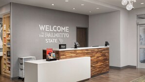 A reception desk with a "Welcome to the Palmetto State" sign, a wooden accent wall, a minibar, and a digital kiosk. - Hampton Inn & Suites Lexington Columbia (Lexington)