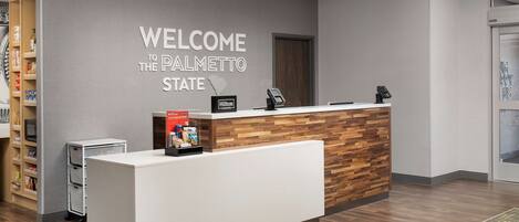 A reception desk with a "Welcome to the Palmetto State" sign, a wooden accent wall, a minibar, and a digital kiosk.