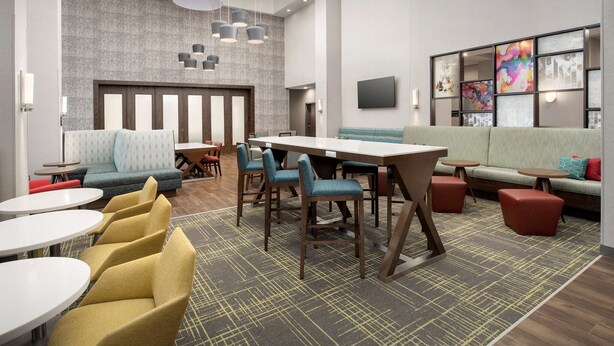 A modern hotel lobby with a large dining table, multiple seating areas, and a large screen wall. - Hampton Inn & Suites Lexington Columbia (Lexington)