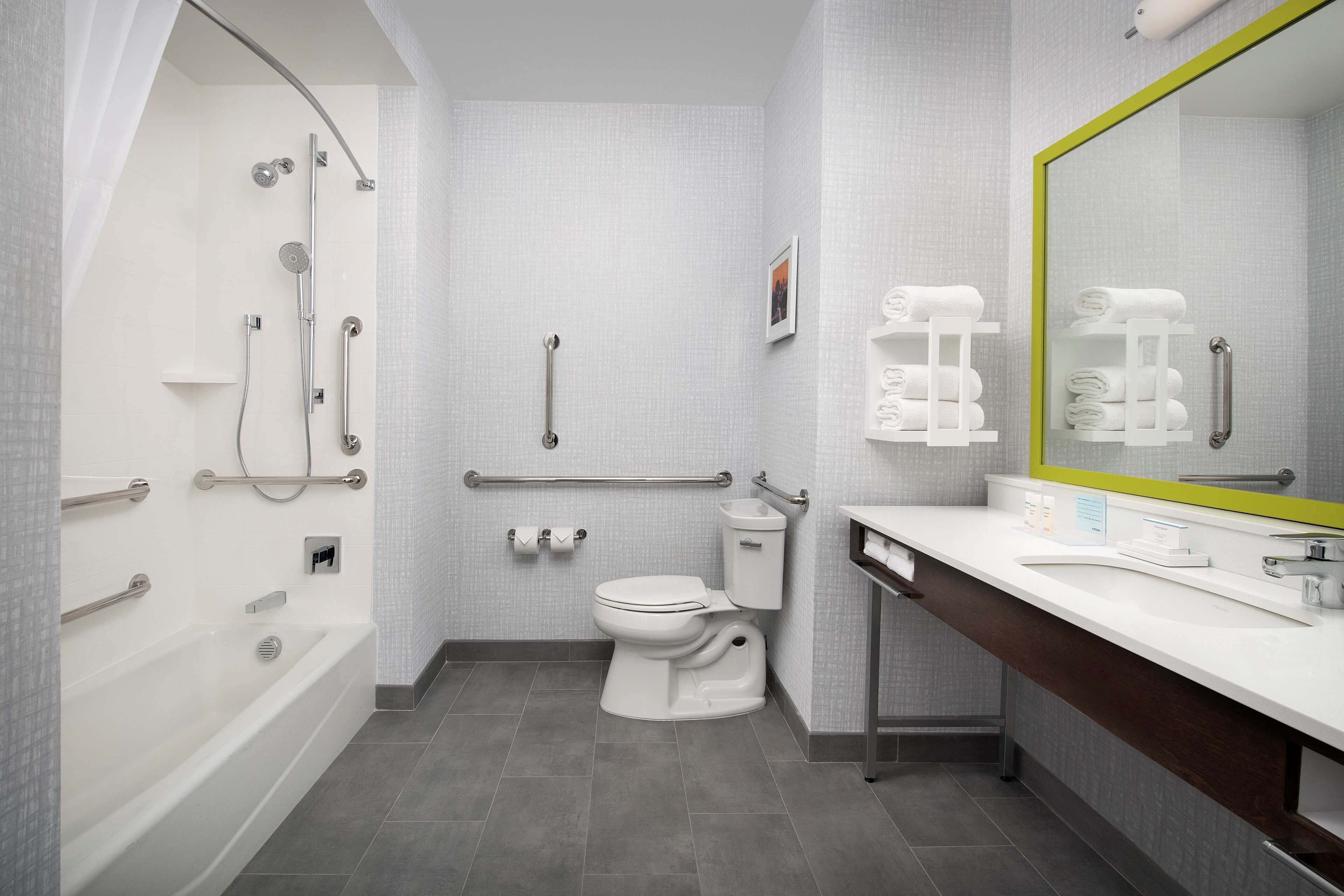 Un baño moderno con bañera, inodoro y lavamanos, equipado con barras de apoyo y una silla de ducha.