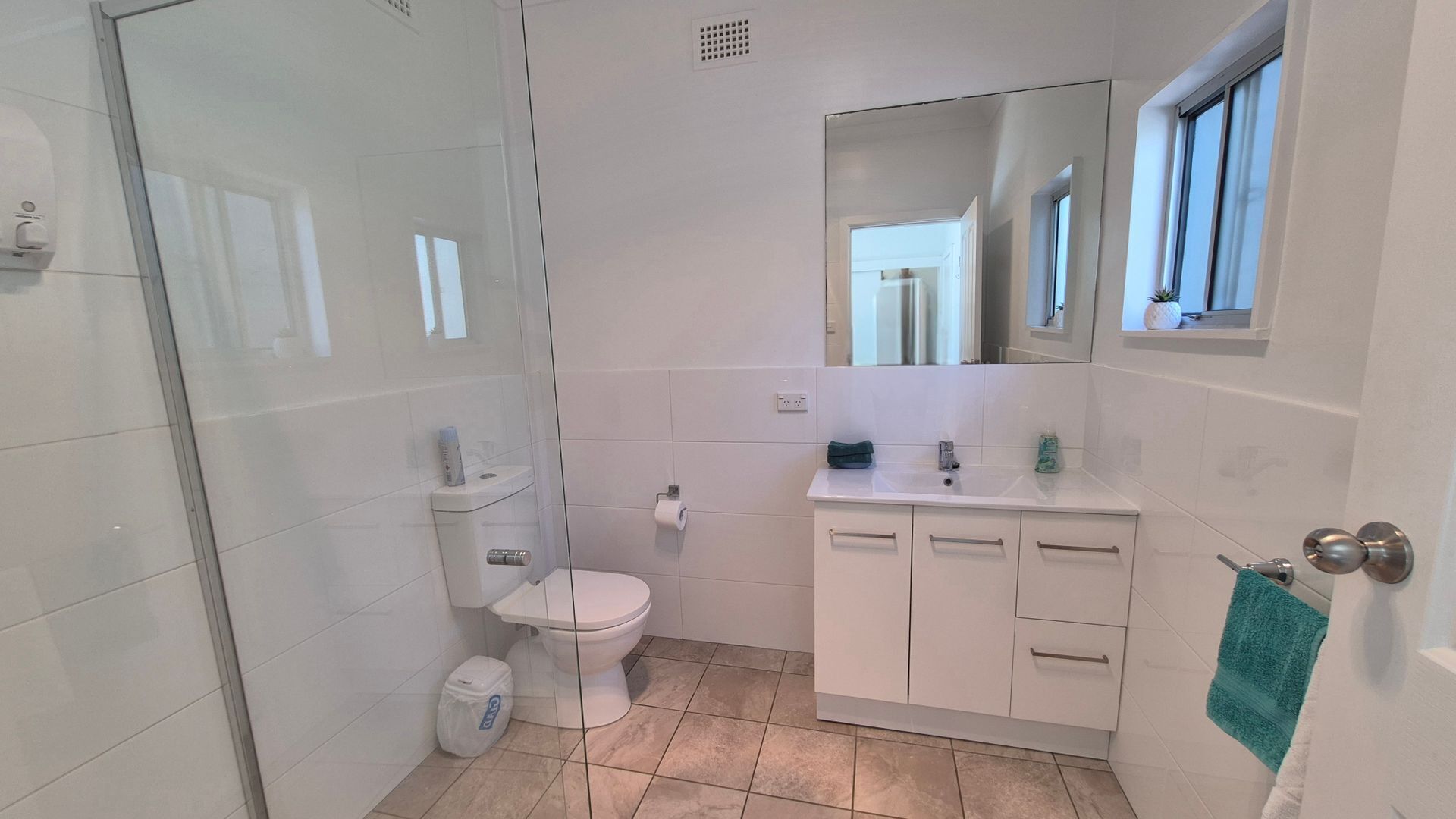 A modern bathroom with a glass shower enclosure, a toilet, a sink with a mirror above, and a towel hanging on the door.