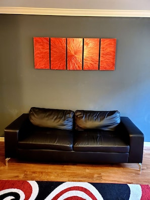 A black leather sofa with two cushions against a dark wall, a red and white patterned rug on the floor, and a set of four red and orange abstract paintings above the sofa.