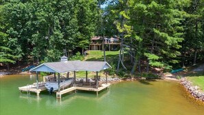 A docked boat house with a covered deck and seating area, surrounded by a lake and dense trees.