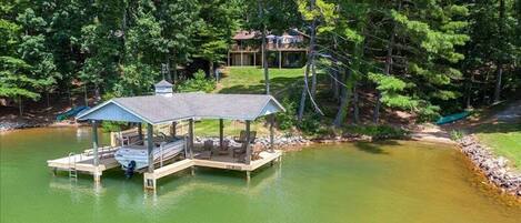 A docked boat house with a covered deck and seating area, surrounded by a lake and dense trees.
