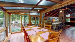 A spacious dining area with a long wooden table and chairs, large windows offering a view of the outdoors, and a kitchen area with a brick wall and modern appliances.