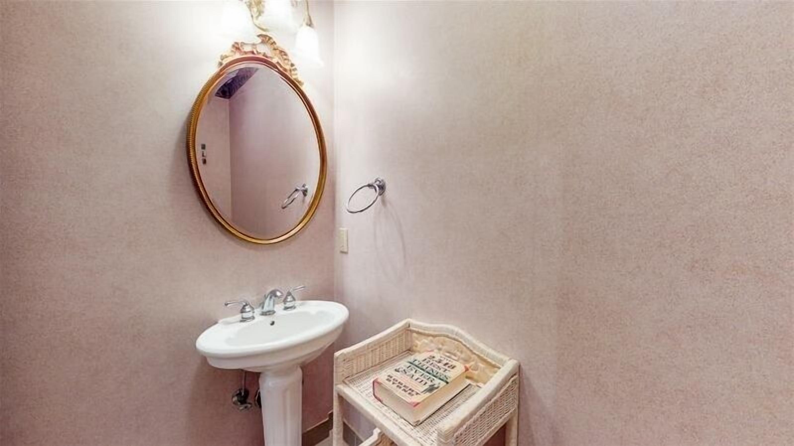 A bathroom with a white sink, a mirror with a gold frame, and a small wicker basket with newspapers.