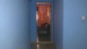 Een smalle gang met blauwe muren en een patroon vloer, die leidt naar een kamer met een rode deur.