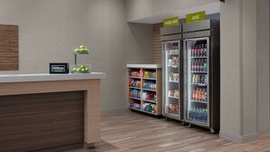 A hotel reception area with a wooden counter, a sign indicating 'Hilton Honors', a 'really cold' sign above a refrigerator, and a 'home' sign above a minibar. - Home2 suites by Hilton, Carlsbad, New Mexico (Carlsbad)