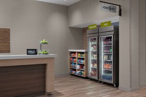 Un área de recepción de hotel con un mostrador de madera, un letrero que indica 'Hilton Honors', un letrero que dice 'muy frío' sobre un refrigerador y un letrero que dice 'hogar' sobre una minibar.