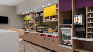 A modern kitchen area with a breakfast bar, a yogurt and milk dispenser, and a variety of food items displayed on shelves. - Home2 suites by Hilton, Carlsbad, New Mexico (Carlsbad)