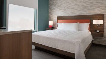 A hotel room with a large bed, a nightstand with a lamp, a desk, and a window with blinds.