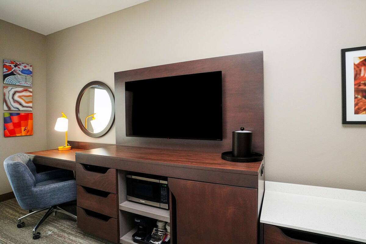 A hotel room with a wooden desk, a flat-screen TV, a round mirror, and a chair.