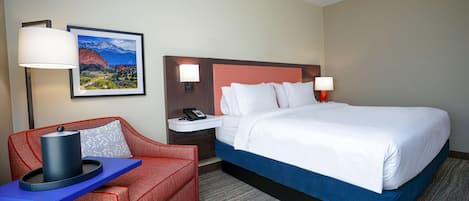 A hotel room with a large bed, a red armchair, a small table with a lamp, and a framed picture on the wall.