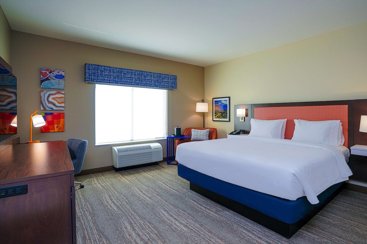 A hotel room with a large bed, a desk, a chair, a window with blinds, and artwork on the walls.