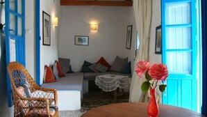Una habitación acogedora con techo de madera, una silla de mimbre, una mesa redonda con un florero y un sofá con cojines, además de una puerta azul.