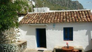 Un edificio blanco con techo de tejas, una puerta azul y una ventana pequeña. Hay una mesa redonda con dos sillas afuera.