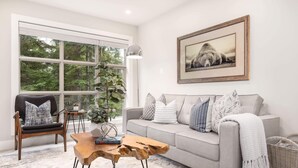 A living room with a grey sofa, wooden coffee table, and a large window with a view of trees. - Peaceful Forest View A/C Pool BBQ Netflix | The Suite Life Whistler at Aspens (Whistler)