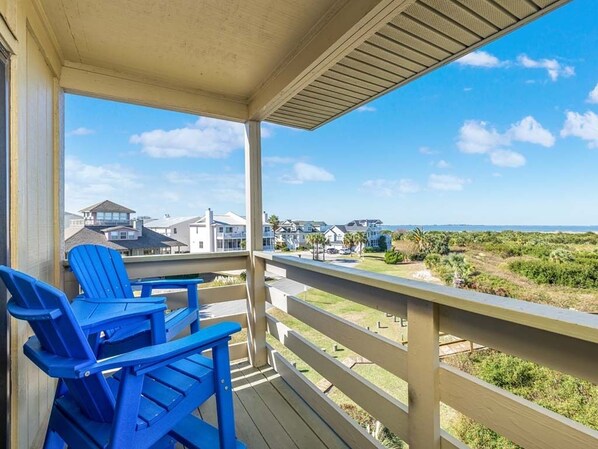 A covered balcony with blue Adirondack chairs, a wooden deck, and a view of houses and the ocean.