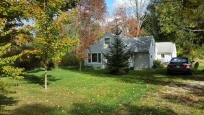 A two-story house with a garage, surrounded by a grassy yard and trees with autumn foliage. - Cozy Cottage close to the Lake (Harbert)