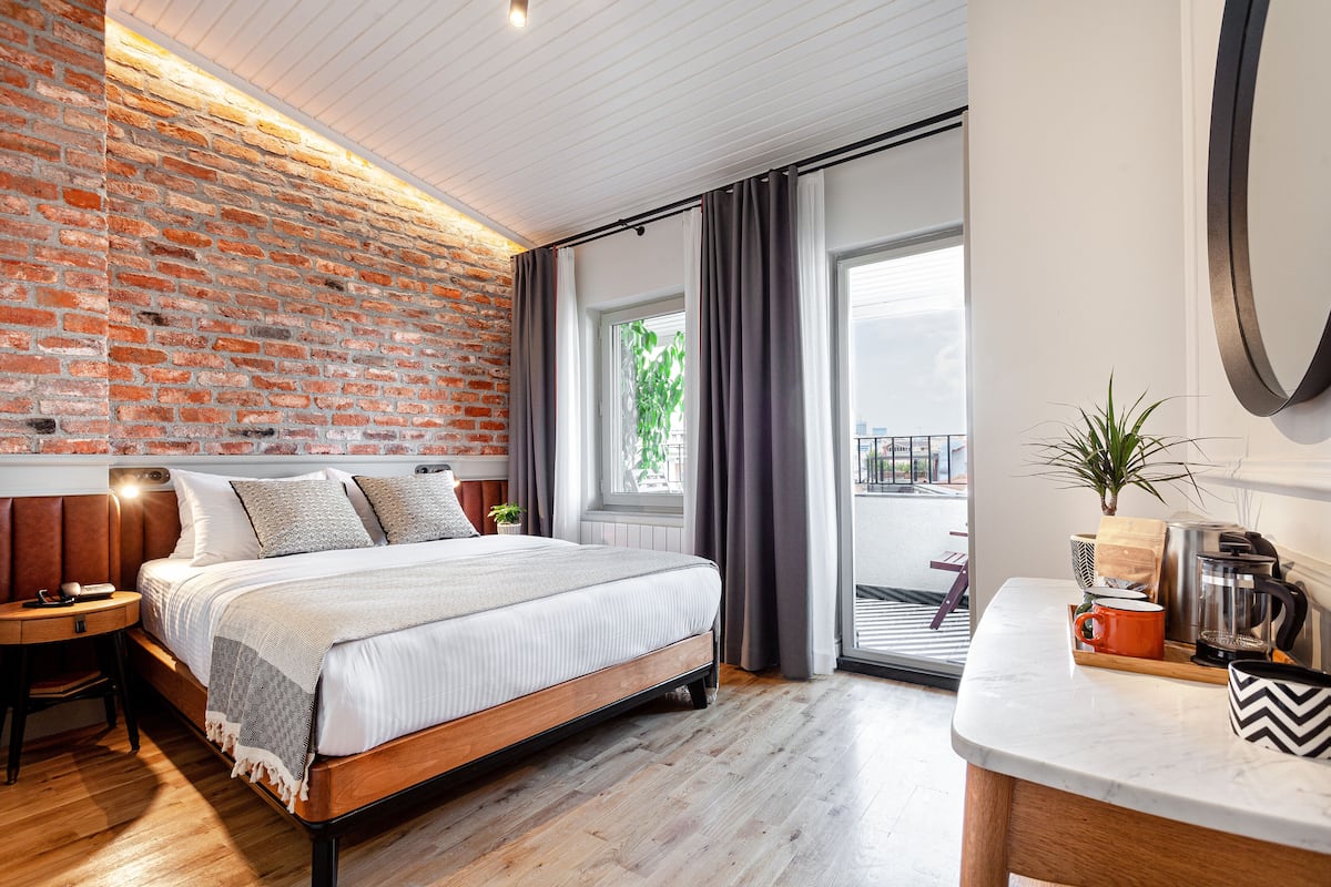 A modern bedroom with a brick wall, a large bed, a wooden nightstand, a desk, and a view of the outdoors.