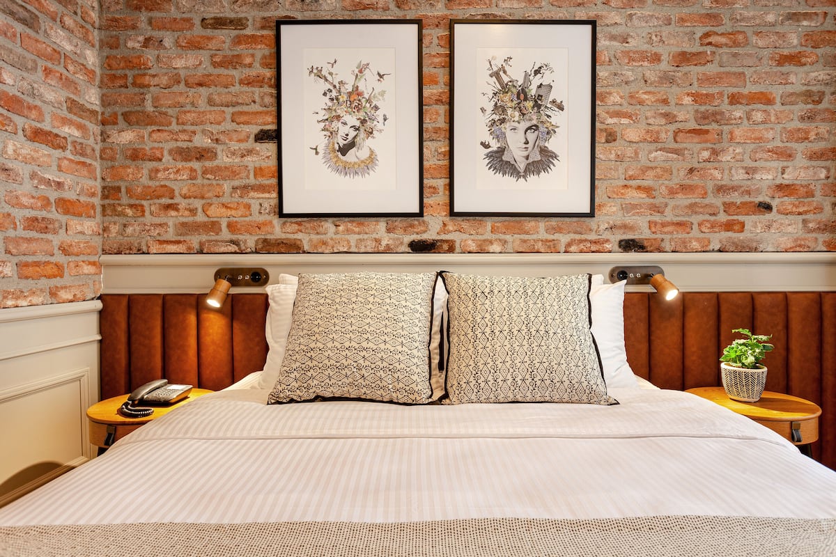 A hotel room with a brick wall, two framed artworks, a bed with patterned pillows, a bedside lamp, a telephone, and a small table with a plant.