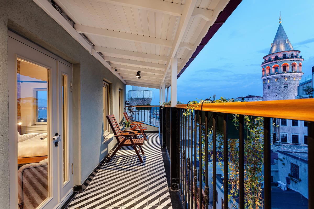 A balcony with a checkered floor, a wooden table, and chairs, overlooking a historic tower and buildings.