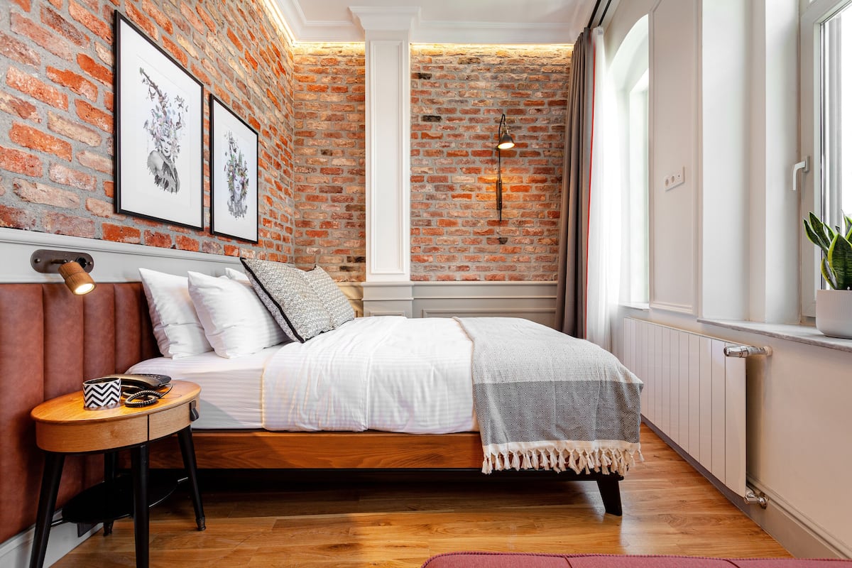 A bedroom with a brick wall, a bed with white bedding, a small wooden table, and a potted plant.