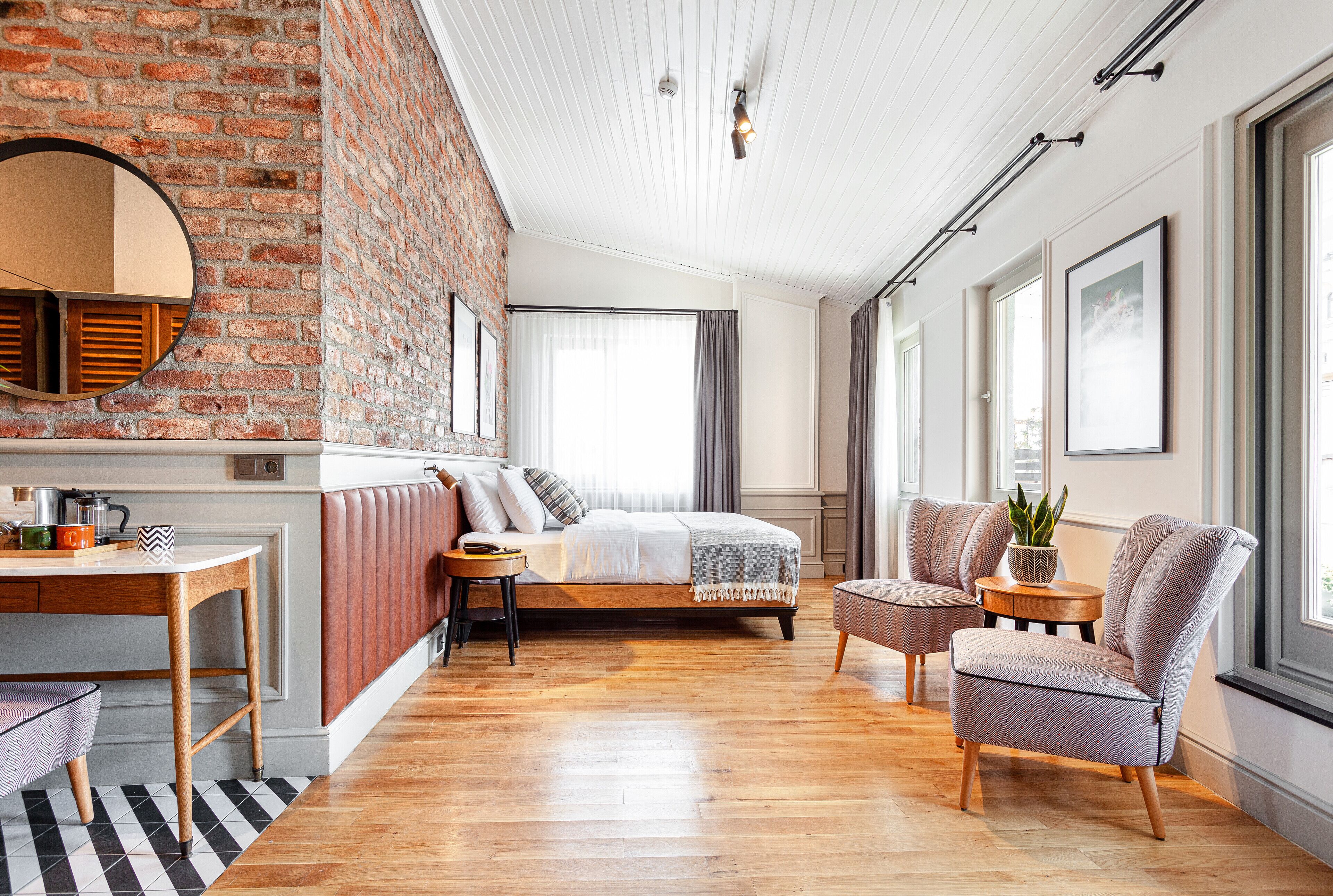 A modern bedroom with a brick wall, wooden flooring, a bed with a headboard, a desk with a chair, and a window with curtains.