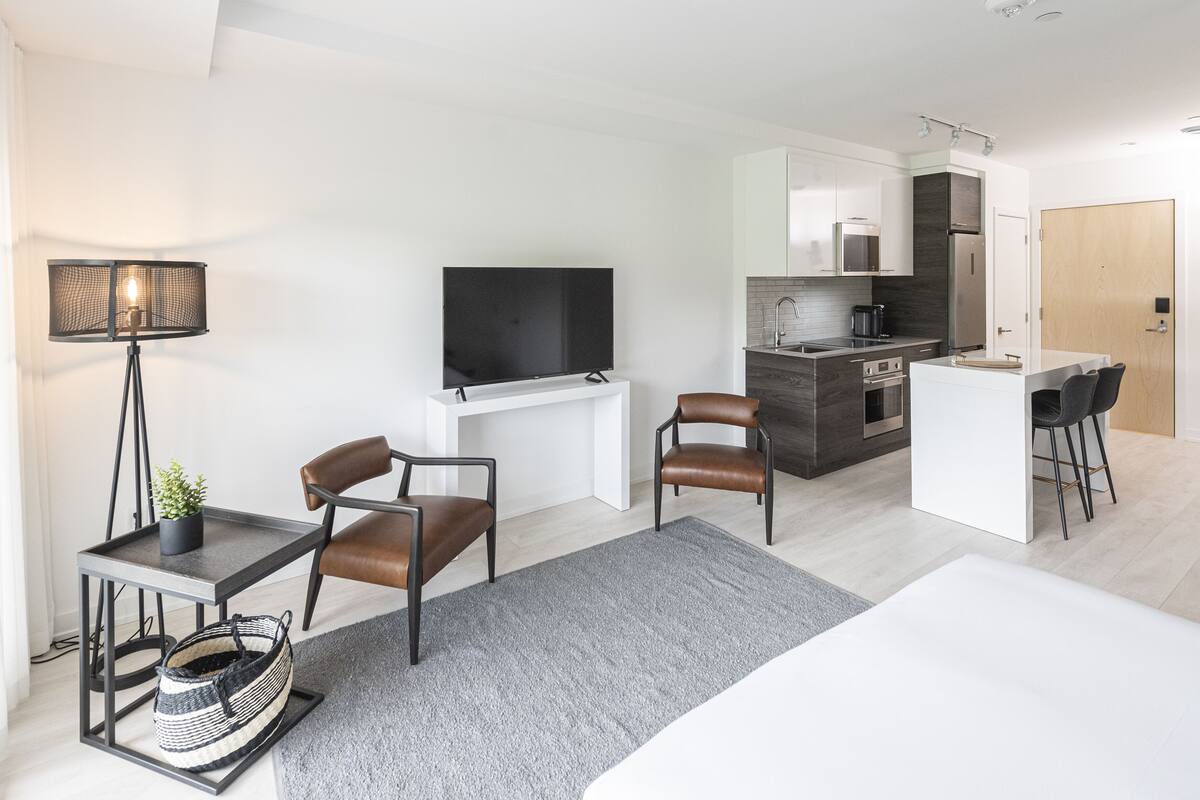 Una habitación moderna y luminosa con televisor de pantalla plana, zona de comedor con mesa y sillas, cocina equipada con refrigerador y estufa, y un ventanal grande.
