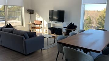 Ein modernes Wohnzimmer mit einer Couch, einem Esstisch, Stühlen, einem Fernseher und großen Fenstern mit Blick auf Bäume.