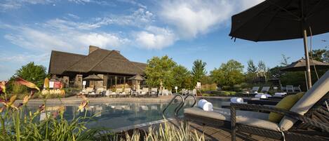 A pool area with lounge chairs and umbrellas, surrounded by greenery and a building with a sloped roof.