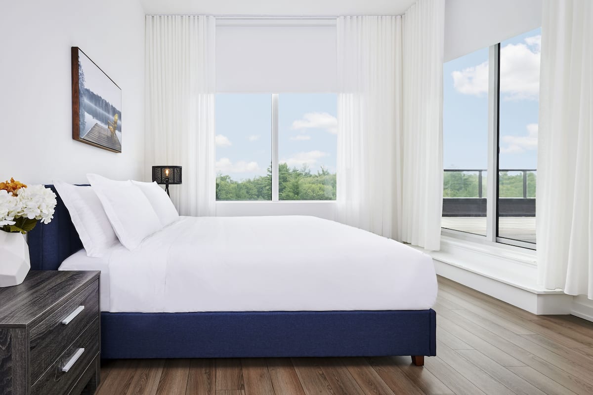 Un dormitorio moderno con una cama grande, ropa de cama blanca, un cabecero azul oscuro, una mesita de noche con un jarrón de flores, un cuadro en la pared y un amplio ventanal con vistas a zonas verdes.
