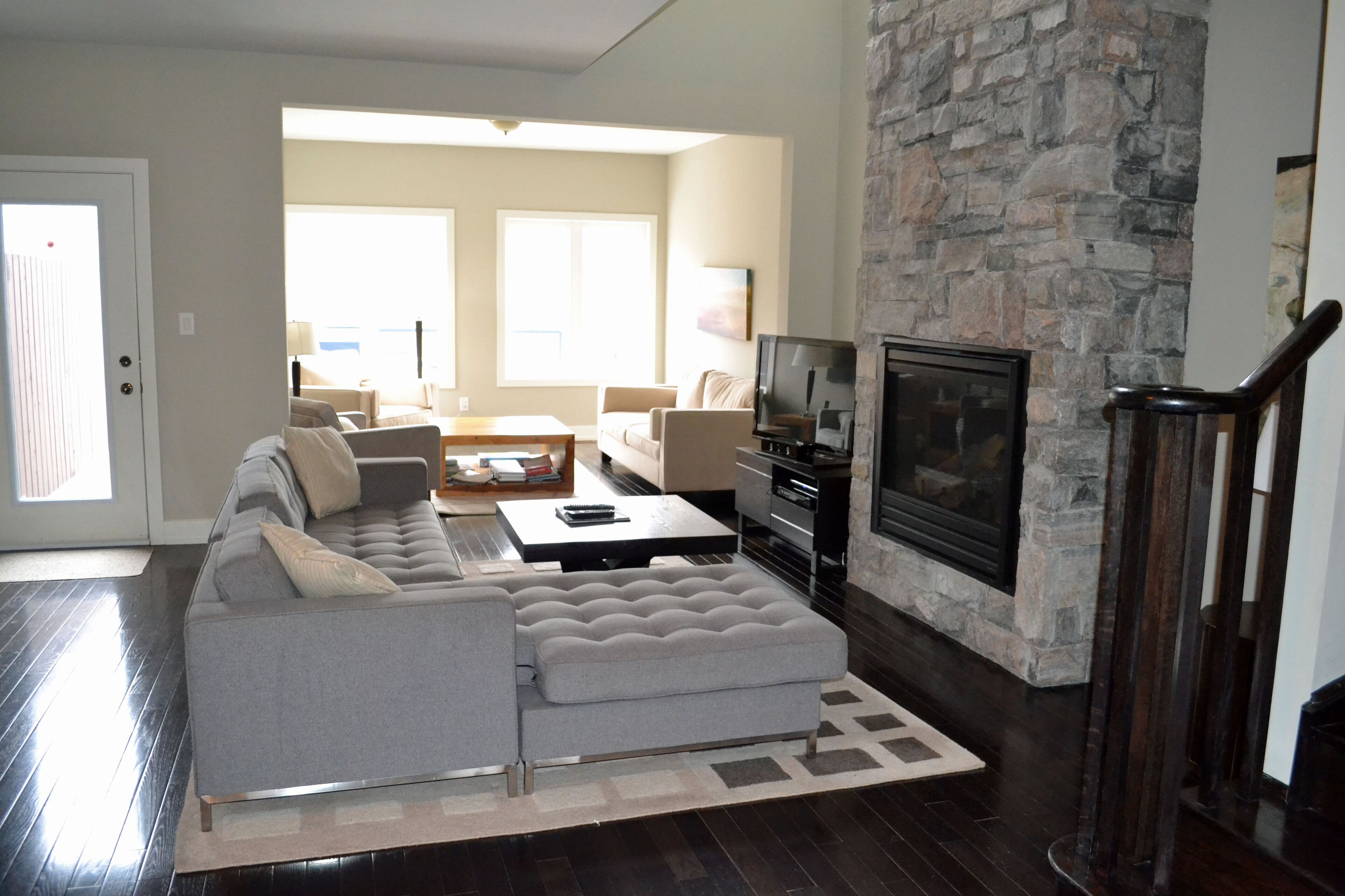 A modern living room with a stone fireplace, a grey sectional sofa, and a flat-screen TV.