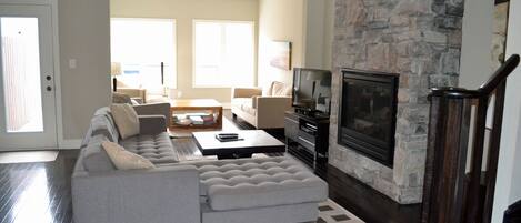 A modern living room with a stone fireplace, a grey sectional sofa, and a flat-screen TV.
